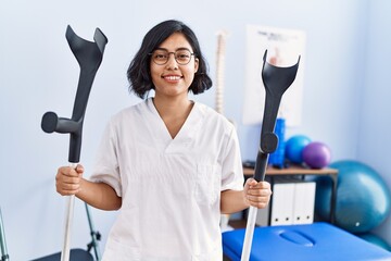 Young latin woman wearing physiotherapist uniform holding crutches at physiotherapy clinic