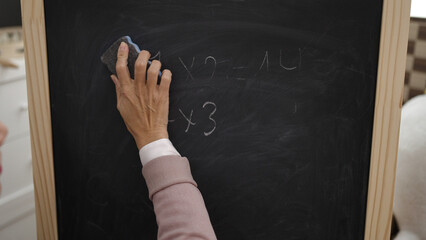 Young beautiful hispanic woman preschool teacher erasing blackboard at kindergarten