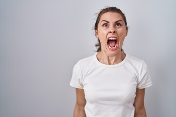 Beautiful brunette woman standing over isolated background angry and mad screaming frustrated and...