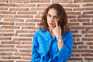 Beautiful brunette woman standing over bricks wall pointing to the eye watching you gesture, suspicious expression