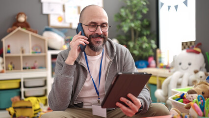 Young bald man preschool teacher using touchpad speaking on the phone at kindergarten