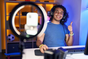 Hispanic man with curly hair playing video games recording with smartphone smiling happy and positive, thumb up doing excellent and approval sign
