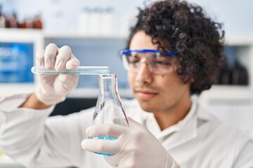 Young hispanic man wearing scientist uniform pouring liquid on test tube at laboratory