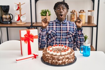 African man with dreadlocks celebrating birthday holding big chocolate cake amazed and surprised looking up and pointing with fingers and raised arms.