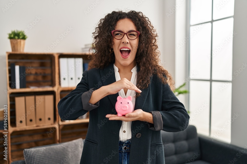 Sticker Young hispanic woman holding piggy bank angry and mad screaming frustrated and furious, shouting with anger. rage and aggressive concept.