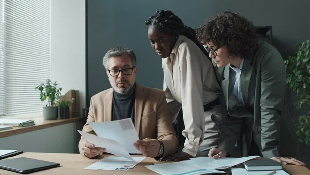 Medium shot of three HRs, Caucasian senior man, Caucasian short-haired woman, young black woman, looking through CVs, discussing applicants, trying to come up with right decision