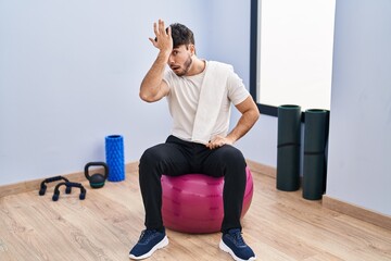 Hispanic man with beard sitting on pilate balls at yoga room surprised with hand on head for mistake, remember error. forgot, bad memory concept.