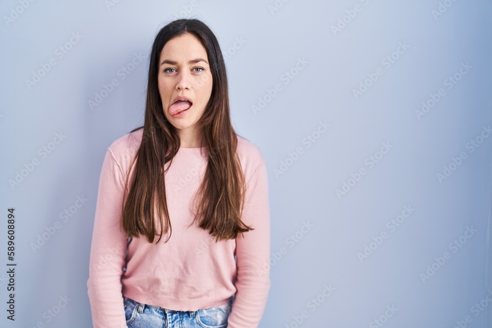 Sticker Young brunette woman standing over blue background sticking tongue out happy with funny expression. emotion concept.