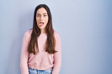Obraz premium Young brunette woman standing over blue background sticking tongue out happy with funny expression. emotion concept.
