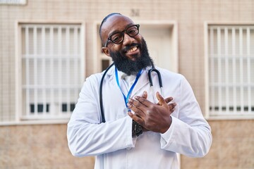 African american man wearing doctor uniform and stethoscope smiling with hands on chest, eyes...