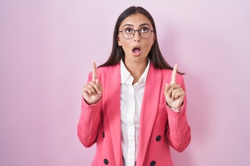 Young hispanic woman wearing business clothes and glasses amazed and surprised looking up and pointing with fingers and raised arms.