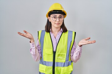 Hispanic girl wearing builder uniform and hardhat clueless and confused expression with arms and hands raised. doubt concept.