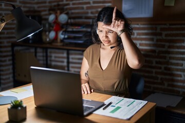 Young hispanic woman working at the office at night making fun of people with fingers on forehead...