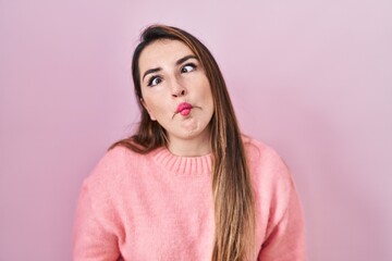 Young hispanic woman standing over pink background making fish face with lips, crazy and comical gesture. funny expression.