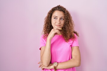 Young caucasian woman standing over pink background with hand on chin thinking about question, pensive expression. smiling and thoughtful face. doubt concept.