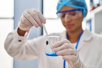 Young latin woman wearing scientist uniform mixing liquid at laboratory
