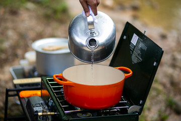 Pouring hot water from a kettle into a red enameled cast iron dutch oven on camping stove