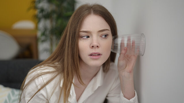 Young blonde woman spying neighbours with glass on wall at home