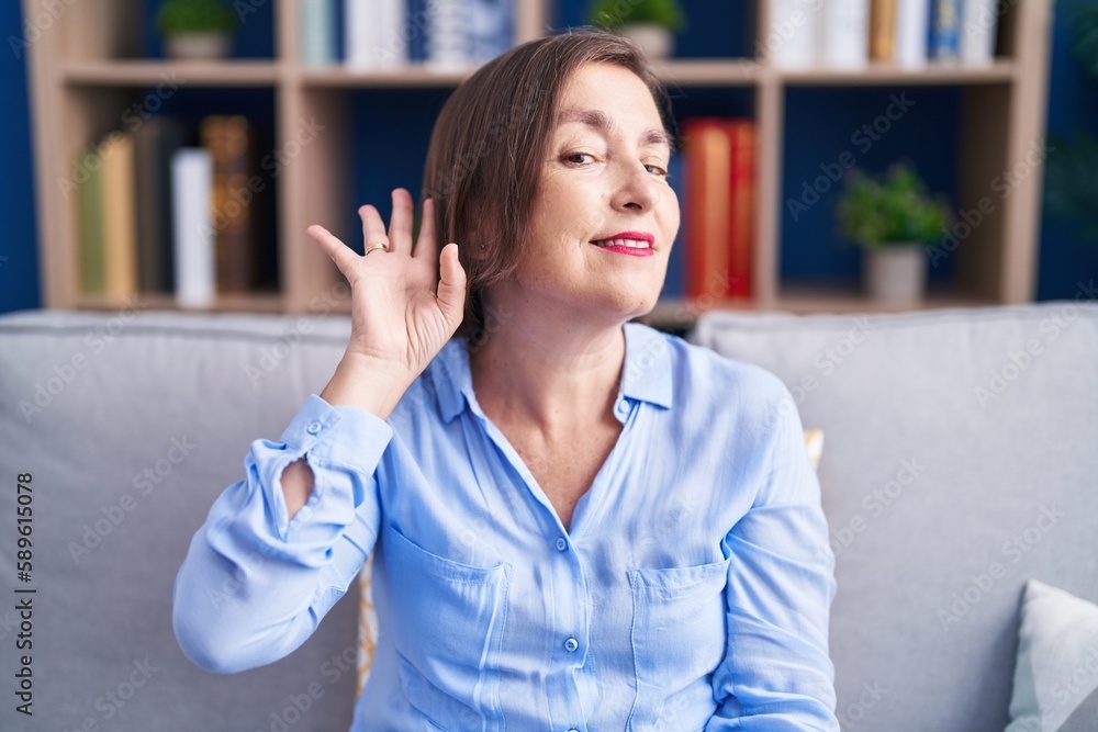 Sticker Middle age hispanic woman sitting on the sofa at home smiling with hand over ear listening an hearing to rumor or gossip. deafness concept.