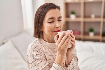 Middle age woman smelling coffee sitting on bed at bedroom