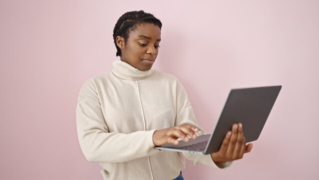 African american woman standing with relaxed expression using laptop over isolated pink background