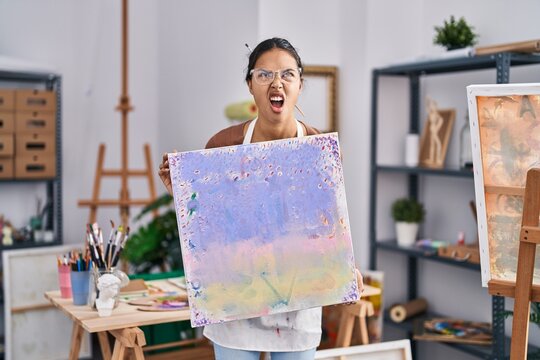 Young brazilian woman holding painter canvas at art studio angry and mad screaming frustrated and furious, shouting with anger looking up.