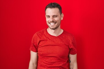 Obraz premium Young hispanic man standing over red background winking looking at the camera with sexy expression, cheerful and happy face.