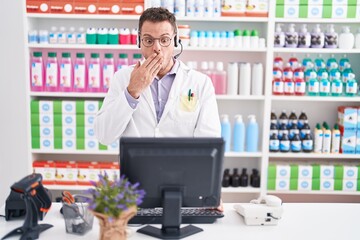 Young hispanic man working at pharmacy drugstore wearing headset covering mouth with hand, shocked and afraid for mistake. surprised expression