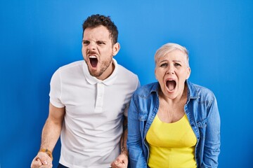 Young brazilian mother and son standing over blue background angry and mad screaming frustrated and furious, shouting with anger. rage and aggressive concept.
