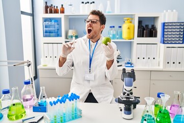 Young hispanic man working at scientist laboratory holding apple angry and mad screaming frustrated and furious, shouting with anger. rage and aggressive concept.
