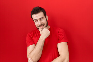 Young hispanic man wearing casual red t shirt looking confident at the camera smiling with crossed arms and hand raised on chin. thinking positive.
