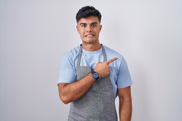 Hispanic young man wearing apron over white background pointing aside worried and nervous with...