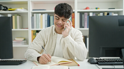 Young hispanic man student talking on smartphone writing notes at library university