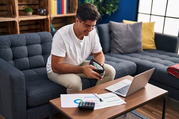Young hispanic man using laptop holding wallet with dollars at home