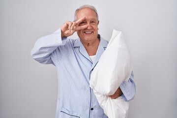 Senior man with grey hair wearing pijama hugging pillow doing peace symbol with fingers over face, smiling cheerful showing victory