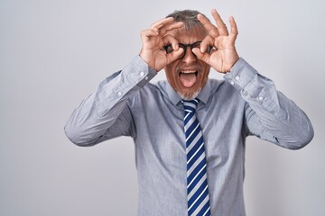 Hispanic business man with grey hair wearing glasses doing ok gesture like binoculars sticking tongue out, eyes looking through fingers. crazy expression.