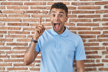 Brazilian young man standing over brick wall pointing finger up with successful idea. exited and happy. number one.