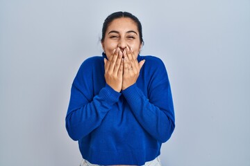 Young hispanic woman standing over isolated background laughing and embarrassed giggle covering...