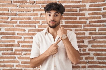 Arab man with beard standing over bricks wall background in hurry pointing to watch time, impatience, looking at the camera with relaxed expression