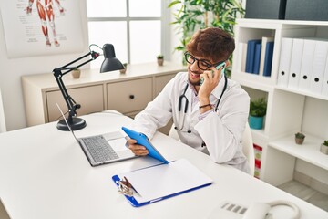 Young arab man wearing doctor uniform talking on smartphone using touchpad at clinic