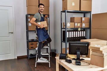 Young hispanic man business worker standing on ladder holding package at storehouse