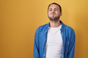 Hispanic man standing over yellow background puffing cheeks with funny face. mouth inflated with air, crazy expression.