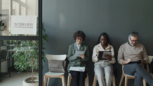 Medium full shot of Caucasian woman, black woman and Caucasian man waiting in line for job interview, looking through their CVs, then taking eyes from them, amiably looking at camera and smiling
