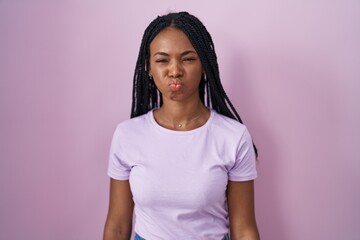Fototapeta premium African american woman with braids standing over pink background puffing cheeks with funny face. mouth inflated with air, crazy expression.