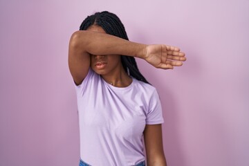 African american woman with braids standing over pink background covering eyes with arm, looking serious and sad. sightless, hiding and rejection concept