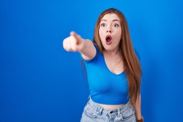 Fototapeta premium Redhead woman standing over blue background pointing with finger surprised ahead, open mouth amazed expression, something on the front