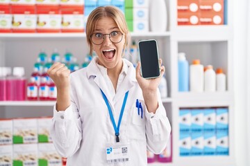 Young caucasian woman working at pharmacy drugstore showing smartphone screen screaming proud, celebrating victory and success very excited with raised arms