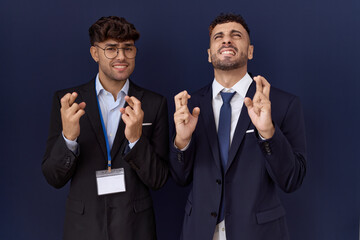 Two hispanic business men wearing business clothes gesturing finger crossed smiling with hope and eyes closed. luck and superstitious concept.