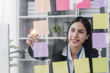 Young businesswoman creative team using post it notes in glass wall to writing strategy business plan to development grow to success.