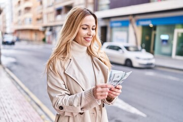 Young blonde woman smiling confident counting dollars at street